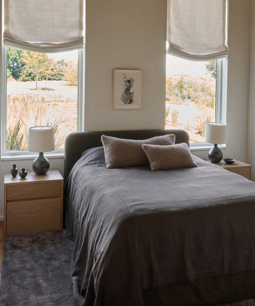 A cozy bedroom features a neatly made bed with a gray blanket, beige pillows, wooden nightstands and matching lamps, plus the Bristol Rug underfoot. Large windows with fabric shades fill the room with natural light.