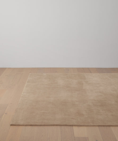 The Bristol Rug, in beige, is handwoven and placed on a light wooden floor in a minimalist room with a plain white wall backdrop.