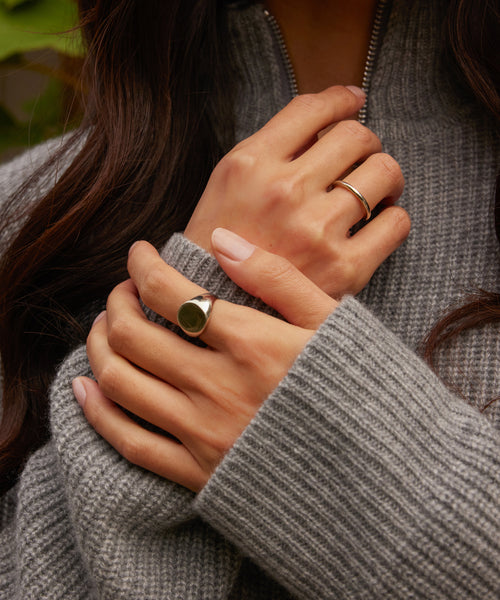 Close-up of hands with neatly manicured nails wearing the Brook Gold Band and a silver band, paired with a grey knitted sweater. The blurred background hints at greenery.