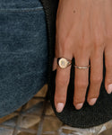 A close-up of a hand in blue jeans rests on herringbone fabric, wearing two rings: a gold signet with a small rectangular gem and the Brook Gold Band—a thin, twisted ring crafted from 14k solid gold.