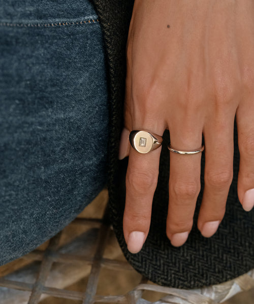 A close-up of a hand in blue jeans rests on herringbone fabric, wearing two rings: a gold signet with a small rectangular gem and the Brook Gold Band—a thin, twisted ring crafted from 14k solid gold.