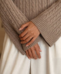 A person in a cozy beige cable-knit Jenni Kayne sweater with flared sleeves, gently clasping their hands over white pants, wears the elegant Brook Gold Band on one finger.