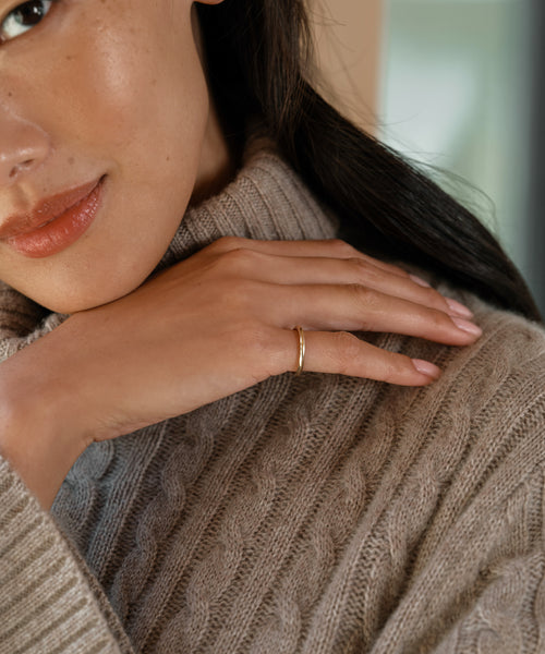 A woman in a beige cable-knit sweater gently rests her hand, showcasing the elegant Brook Gold Band in 14k gold, on her shoulder. Only the lower half of her face is visible, highlighting her smooth skin and subtle makeup.