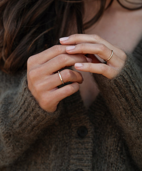 A person wearing a fuzzy brown cardigan gently touches their fingers, showcasing the Brook Gold Band—a simple 14k gold ring. Their long brown hair peeks into view, enhancing the elegant, timeless heirloom style.