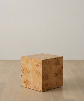 The Sur Side Table is a square wooden cube with a light, natural finish and visible wood grain, evoking a vintage look. It sits on a light wood floor against a plain white wall.
