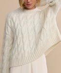 A woman poses with one arm raised, highlighting the upper body and details of the Cable Serena Crewneck—a classic ivory sweater featuring cable-stitching, a ribbed collar, and hem.