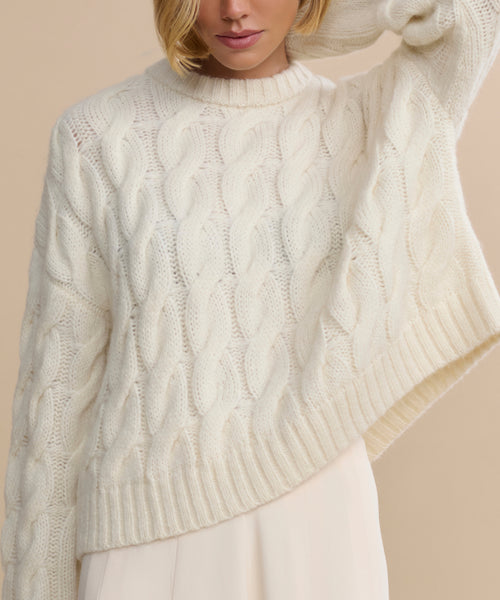 A woman poses with one arm raised, highlighting the upper body and details of the Cable Serena Crewneck—a classic ivory sweater featuring cable-stitching, a ribbed collar, and hem.