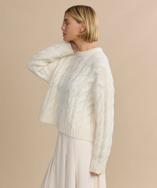 A woman with short blonde hair wears the Cable Serena Crewneck sweater in ivory and a long, light skirt. She stands sideways against a beige background, one hand near her neck.