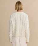 A person with short blonde hair is seen from behind, wearing the Cable Serena Crewneck in ivory and a light pleated skirt, standing against a beige background.
