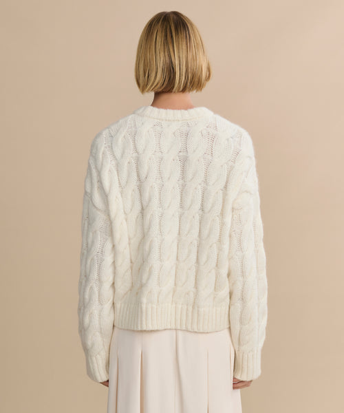 A person with short blonde hair is seen from behind, wearing the Cable Serena Crewneck in ivory and a light pleated skirt, standing against a beige background.