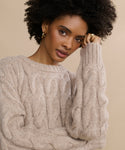 A person with curly hair wearing gold hoop earrings and the Cable Serena Crewneck, a classic cable-stitch alpaca-wool sweater, stands against a neutral background, calmly resting their cheek on their hand and looking at the camera.