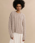 A woman with curly hair models the Cable Serena Crewneck, a loose beige cable-knit sweater, paired with a cream skirt, standing against a plain beige background.
