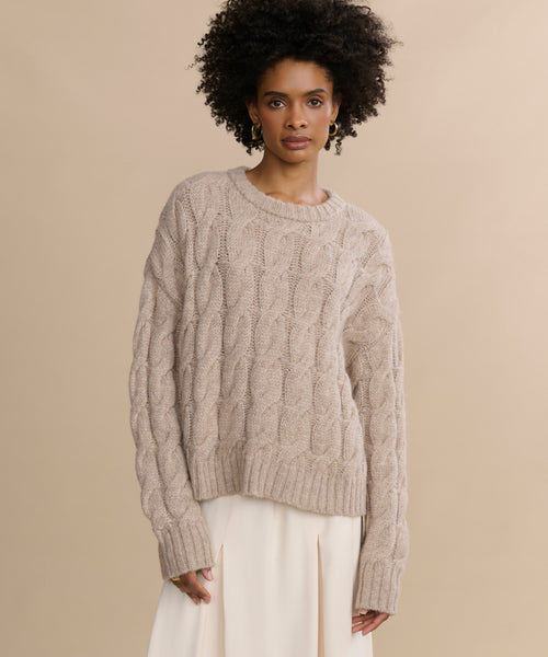 A woman with curly hair models the Cable Serena Crewneck, a loose beige cable-knit sweater, paired with a cream skirt, standing against a plain beige background.