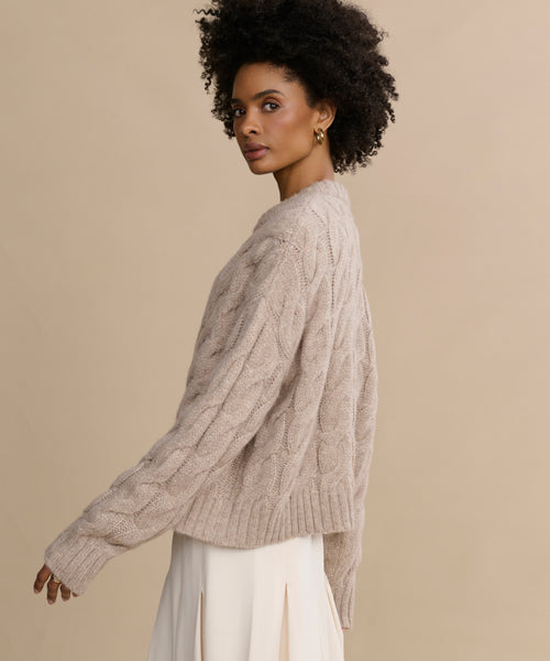 Woman with curly hair wearing the Cable Serena Crewneck sweater and an off-white skirt, standing sideways against a plain beige background.