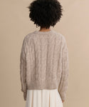 A person with short, curly hair is seen from behind wearing the Cable Serena Crewneck—a chunky beige alpaca-wool sweater—with a cream skirt, all set against a plain beige background.