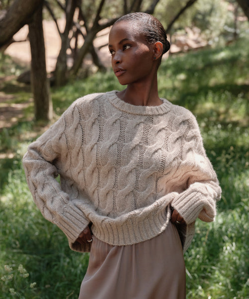 A woman stands in a wooded area, sunlight filtering through the trees, wearing the Cable Serena Crewneck sweater in cream and a light beige skirt. She looks to the side with her hands on her sweater’s hem.