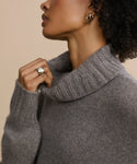 A woman with short, curly hair, silver hoop earrings, and a Campbell Cigar Band ring lifts the collar of her soft gray turtleneck sweater near her chin. Her face is partially out of frame.