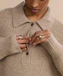 A person wearing a chunky beige knit sweater with a collar fastens a button. Silver rings, including the Campbell Cigar Band, adorn their hands. The focus is on their hands and sweater against a neutral beige background.