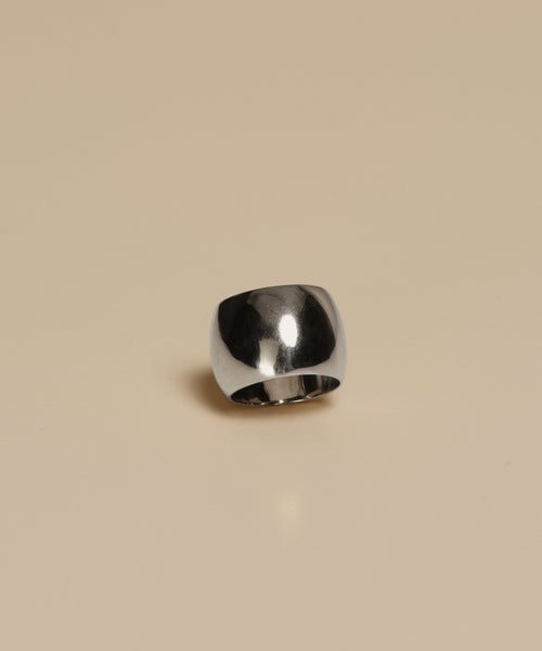 The Campbell Cigar Band, a wide and polished ring, stands upright on a light beige surface, reflecting its surroundings—an elegant piece from Jenni Kayne Jewelry.