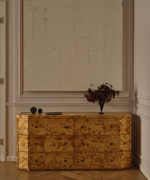 The Carbon Dresser, featuring six soft-close drawers in golden burlwood, stands against the wall. It's topped with a black vase of dark red leaves and two cups, while a large textured monochromatic white artwork hangs above.