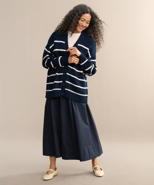 A woman with long curly hair smiles in the Chloe Cardigan, styled with a navy midi skirt, light top, and cream shoes against a beige background—an ideal warm-weather sweater look.