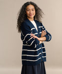 A woman with long, curly gray hair poses confidently against a neutral background, arms crossed, wearing the Chloe Cardigan in Navy/Ivory Stripe over a white top and dark skirt—a perfect warm-weather sweater choice.