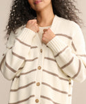A person with curly hair wears the Chloe Cardigan, an ivory taupe striped sweater featuring brown horizontal lines and buttons. They hold it closed at the chest, their face partially out of frame—ideal for warm-weather layering.