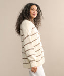 A woman with long, curly hair models the Chloe Cardigan in ivory taupe stripe, standing sideways and gazing at the camera against a beige background—an ideal choice for warm-weather style.