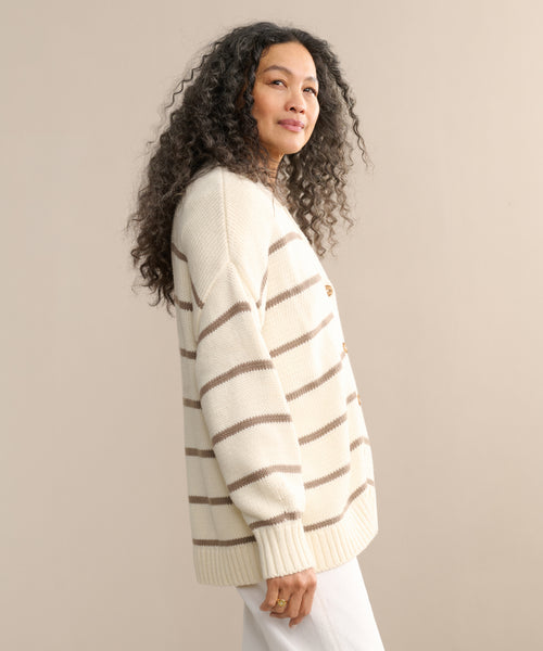A woman with long, curly hair models the Chloe Cardigan in ivory taupe stripe, standing sideways and gazing at the camera against a beige background—an ideal choice for warm-weather style.