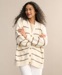 A woman with long curly hair wears the Chloe Cardigan in ivory taupe stripes over a beige top and white pants. With her arms crossed and smiling to the side, she shows how this cardigan brings effortless style to warm-weather looks.
