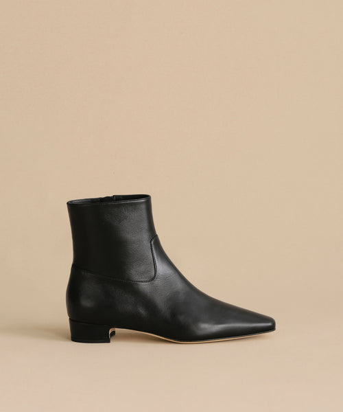 The Leather Carmine Boot, a single black leather boot with a pointed toe and low heel, is shown against a beige background, highlighting its classic bootie style.