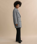 A woman with curly hair and a relaxed expression stands sideways against a beige background, wearing the Caroline Coat—an oversized gray double-breasted style—paired with black pants and black shoes.