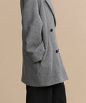 A person wears the Caroline Coat—an oversized gray wool coat with black buttons—and black pants, posing against a plain beige background. Only their torso and upper legs are visible.
