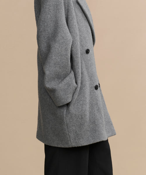 A person wears the Caroline Coat—an oversized gray wool coat with black buttons—and black pants, posing against a plain beige background. Only their torso and upper legs are visible.