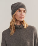 A blonde woman wears the Cashmere Alpine Beanie and a matching gray fisherman rib turtleneck sweater, looking slightly to the side against a plain, neutral background.