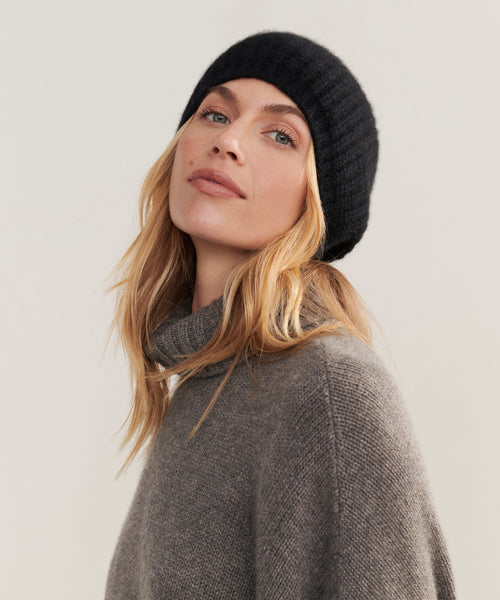 A woman with wavy blonde hair wears the Cashmere Alpine Beanie in black and a gray turtleneck sweater, posing against a plain light background with a neutral expression—showcasing this cozy winter essential.