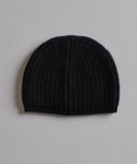 The Cashmere Alpine Beanie, in classic black ribbed knit, is displayed flat on a light gray textured surface—an essential winter accessory.
