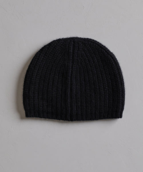 The Cashmere Alpine Beanie, in classic black ribbed knit, is displayed flat on a light gray textured surface—an essential winter accessory.