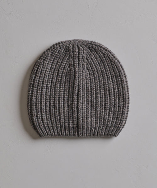The Cashmere Alpine Beanie, featuring a classic fisherman rib texture in gray, is displayed flat on a light, textured surface.
