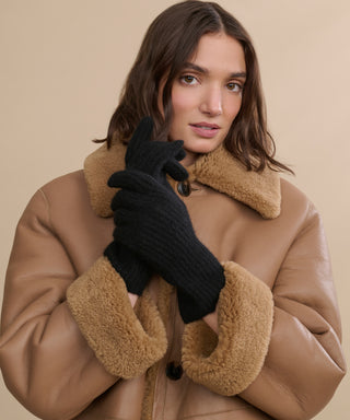 A woman wearing a brown shearling coat and Cashmere Alpine Gloves stands against a beige background, looking at the camera with her gloved hands near her face, highlighting chic winter style.