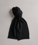 The Cashmere Alpine Scarf in black, featuring a ribbed knit, is displayed on a light gray surface, with one end casually looped to form a knot.