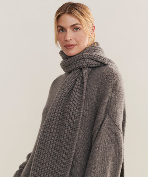 A woman with blonde hair in a low ponytail wears the Cashmere Alpine Scarf—a chunky gray knit winter accessory—along with a matching gray sweater, looking at the camera with a soft, neutral expression against a light background.