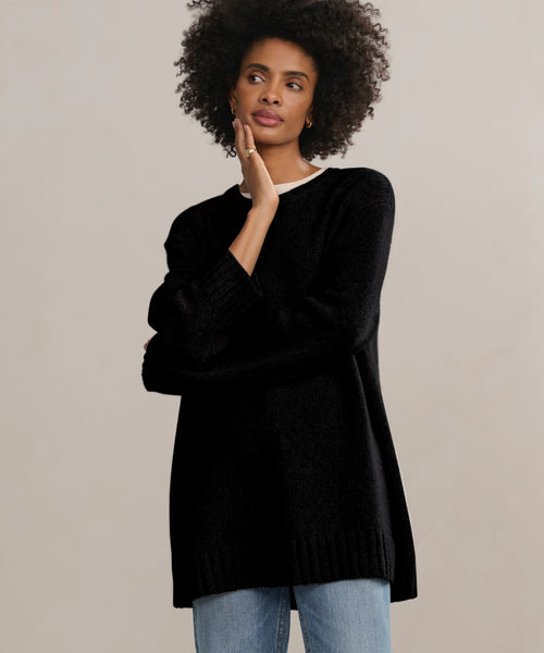 A woman with curly hair wears the Cashmere Amelia Crewneck in black over a white top and blue jeans, standing against a plain light background and thoughtfully touching her face with one hand.