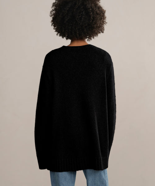 A person with curly hair, seen from behind against a plain light background, wears the Cashmere Amelia Crewneck in black paired with blue jeans.