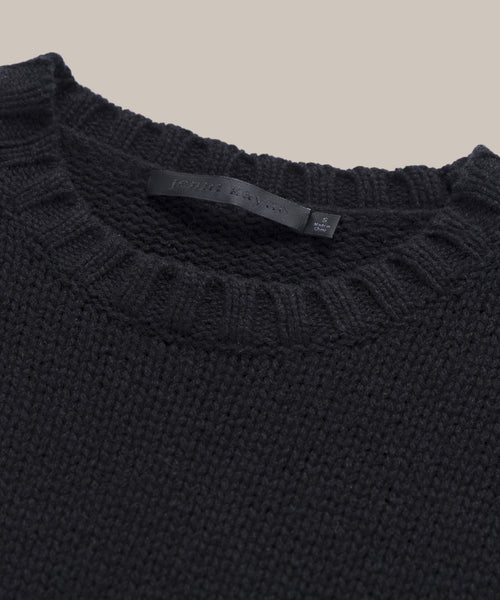 Close-up of the Cashmere Amelia Crewneck in black, featuring a ribbed crew neckline and an inner fabric label. Shown against a plain light beige background.