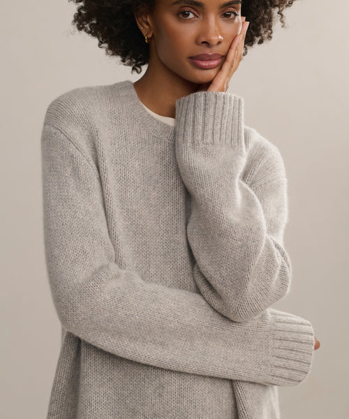 A woman with curly hair wears the Cashmere Amelia Crewneck in light gray, standing against a neutral background with one hand touching her face and gazing thoughtfully at the camera.