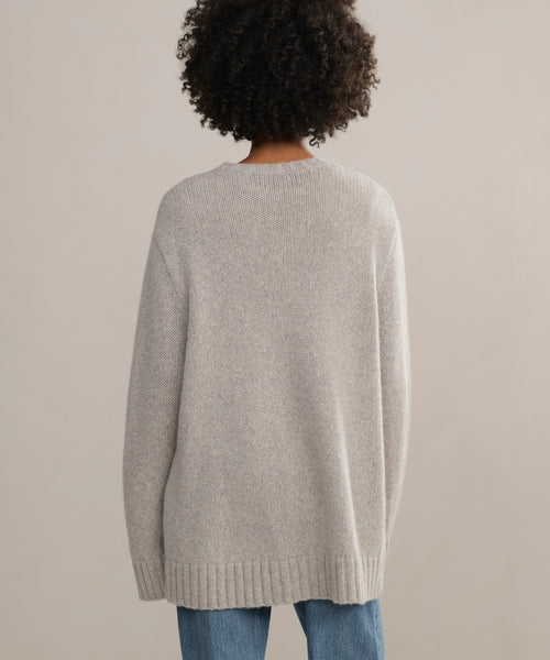 A person with short curly hair, seen from behind, is wearing the Cashmere Amelia Crewneck in light gray with blue jeans against a plain light background.