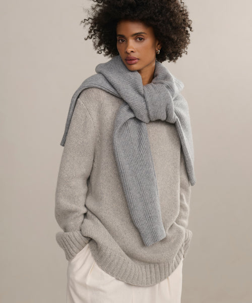 A woman with curly hair wears the Cashmere Amelia Crewneck in gray with a matching oversized cashmere sweater draped over her shoulders and white pants, posing against a plain beige background.