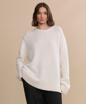 A woman with wavy brown hair wears the oversized ivory Cashmere Amelia Crewneck sweater and loose black pants, standing against a plain beige background.