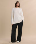 A woman stands against a beige background wearing the Cashmere Amelia Crewneck, paired with wide-leg black pants and white shoes, with one hand in her pocket and a relaxed expression.